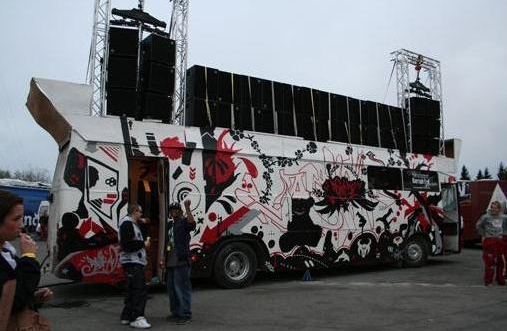 A "russ bus" from 2006. The bus is a white color, painted with a pattern in red and black, reminiscent of a graffiti mural. On the roof of the bus are huge speakers lining the bus from front to back.