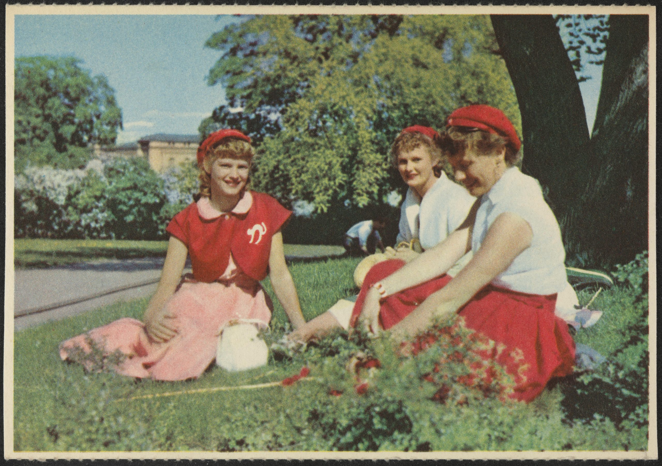 An image of three girls sitting in a park, on the grass. They are wearing red "russ hats" (russeluer). They are also wearing red skirts or red shirts, with the poodle-style skirts dating the picture to around the 1950's.